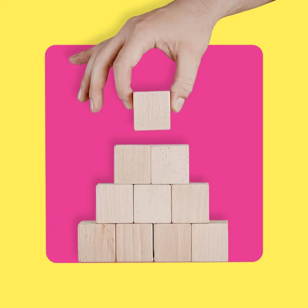 Hand placing a wooden block on top of a pyramid made of blocks, with a pink background and yellow border.