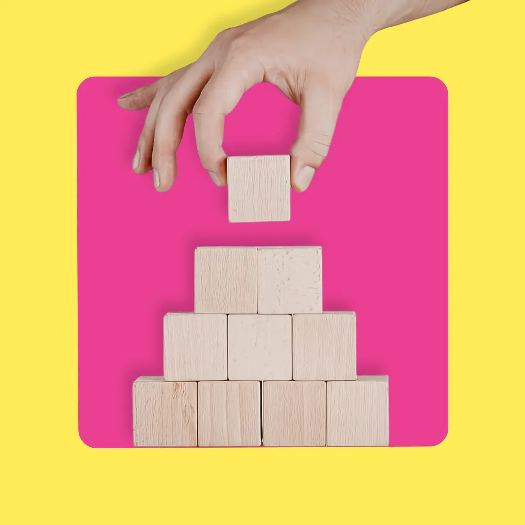 Hand placing a wooden block on top of a pyramid made of blocks, with a pink background and yellow border.